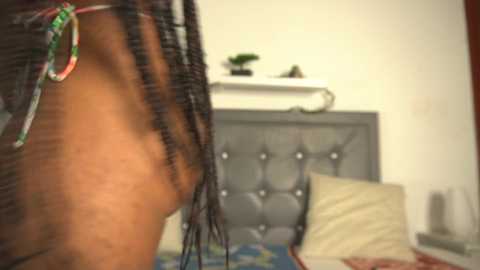 Media: A video of a brown-skinned woman with dreadlocks, wearing colorful bracelets, leaning against a tufted gray headboard in a dimly lit bedroom with a white wall and a potted plant.