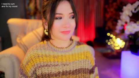 Media: Video of a young woman with dark hair, wearing a beige and yellow striped knit sweater, sitting indoors with warm lighting, blurred background, and text overlay.