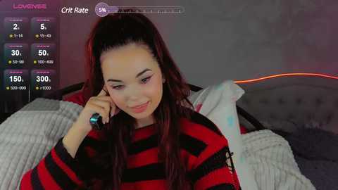 Media: A video of a young woman with long, dark hair in a ponytail, wearing a red and black striped sweater, holding a phone to her ear, sitting on a bed with white and grey blankets.