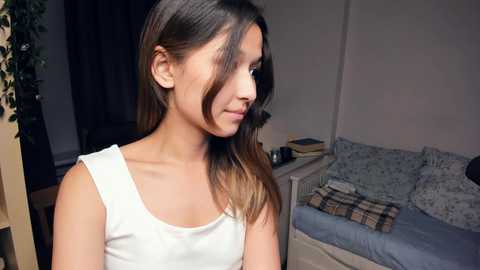 Media: Video of a young woman with long brown hair, wearing a white tank top, standing in a simple bedroom with a bed, plant, and shelves.