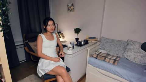 Media: Video of an Asian woman with short black hair, wearing a white sleeveless dress, sitting on a chair in a small, cozy bedroom with a bed, desk, and plants.