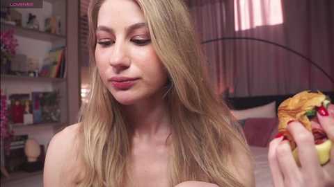 Media: Video of a fair-skinned, blonde woman with straight hair, wearing a strapless top, eating a burger with a lemon slice, in a softly lit, pink-hued bedroom.