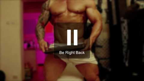 Media: Video of a muscular man with tattoos, wearing white underwear, standing in a dimly lit bedroom with a pink glow and blurred background. Text overlay reads \"Be Right Back.\