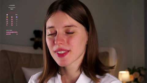 Media: A video of a Caucasian woman with straight brown hair, fair skin, and closed eyes, smiling. She's wearing a white shirt, and the background shows a blurred bedroom with a bed and a lit candle.