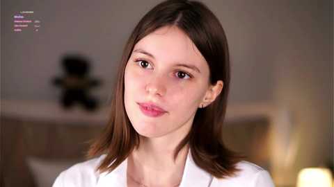 Media: Video of a fair-skinned, brunette woman with straight hair, wearing a white blouse, smiling slightly. Background shows a blurred, beige-toned room with a stuffed bear.