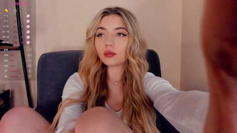 Media: A video of a blonde woman with long wavy hair, wearing a white top, sitting in a dark chair, looking at the camera with a neutral expression.