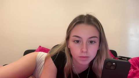 Media: Video of a young Caucasian woman with light skin and long, straight brown hair, wearing a black tank top and white panties, lying on a bed with pink sheets.