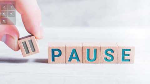 Media: A video of a hand holding a wooden block with the word \"Pause\" in blue letters, placed on a light-colored, textured surface. The background is blurred, with a digital clock visible on the left.