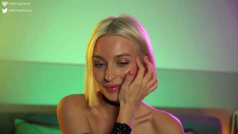 Media: Video of a blonde woman with fair skin, wearing a black choker, touching her face with a soft smile, in a room with green and purple lighting.