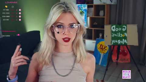 Media: Video of a fair-skinned, blonde woman with large glasses and red lipstick, wearing a beige top, in a modern, artsy room with abstract paintings and a \"Good vibes only\" sign.