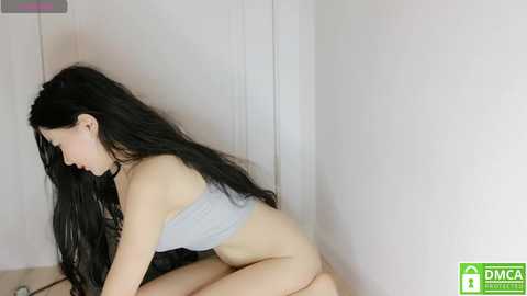 Media: A video of a slender, long-haired Asian woman with pale skin, wearing a light gray bra, sitting on a beige floor against a white wall. She is looking down, with a neutral expression.