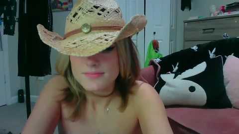 Media: A video of a topless young woman with shoulder-length blonde hair, wearing a straw hat, sitting on a bed with black and white pillows in a cluttered bedroom.