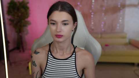 Media: Video of a slender, light-skinned woman with long brown hair, wearing a black-and-white striped top, sitting in a white gaming chair against a pink backdrop.