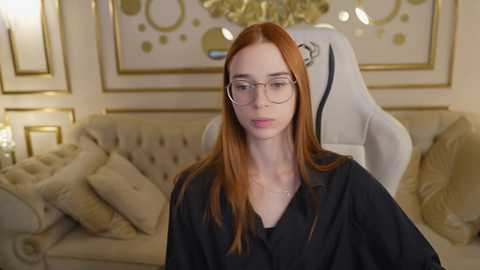 Media: Video of a pale-skinned, red-haired woman with glasses, wearing a black robe, seated in a plush, beige tufted sofa. Background features a gold-framed mirror and ornate wall decor.