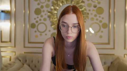 Media: Video of a fair-skinned, red-haired young woman with glasses, wearing a black lace bra, sitting on a beige, tufted sofa. Behind her is a decorative, gold-framed, circular-patterned wall art in an opulent room.