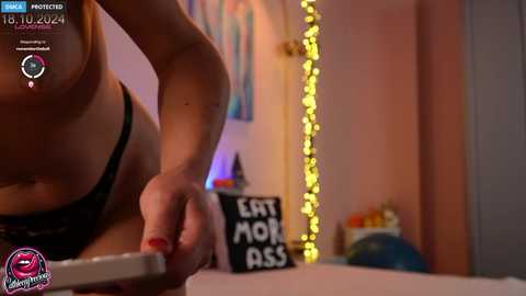 Media: Video of a woman in a black bra, leaning over a table, holding a phone, in a cozy room with fairy lights and \"Eat Mom Ass\" sign.