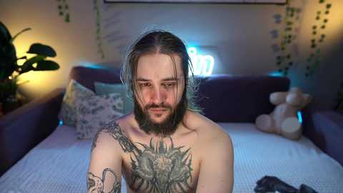 Media: Video of a shirtless, bearded man with long, wet hair, tattoos on chest and arm, sitting on a bed in a dimly lit room with green vines, a potted plant, and stuffed toys.