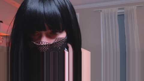 Media: A video of a person with long, straight black hair, wearing a black lace mask covering the eyes, standing in a softly lit room with beige walls and white curtains.