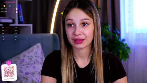 Media: Video of a young woman with straight, shoulder-length blonde hair and light skin, wearing a black top, smiling softly. Background features a modern living room with a gray couch and green curtains.