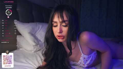 Media: A video of a young woman with long black hair and fair skin, wearing a white lace bra, lying on a bed in a dimly lit room, eyes closed, mouth slightly open, displaying an expression of pleasure.