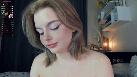 Media: A video of a young, light-skinned woman with shoulder-length blonde hair, wearing minimal makeup, including blue eyeshadow and pink lipstick, standing in a dimly lit bedroom with a bed, a shelf, and a lamp in the background.