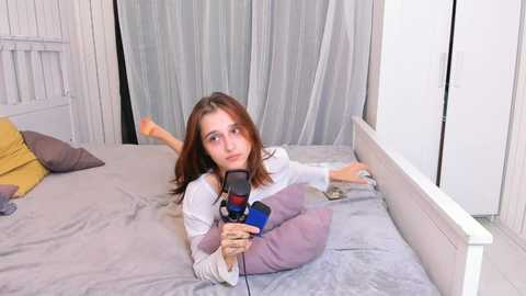 Media: Video of a young Caucasian woman with shoulder-length brown hair, lying on a grey bed with white walls and a white wardrobe, holding a blue microphone, legs bent and crossed.