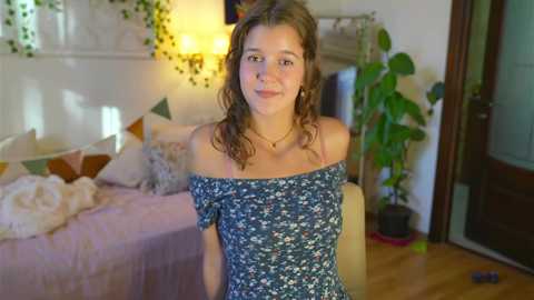 Media: Video of a young woman with curly brown hair, wearing a floral off-shoulder top, sitting in a cozy bedroom with a cream-colored bed, plants, and a wooden door.