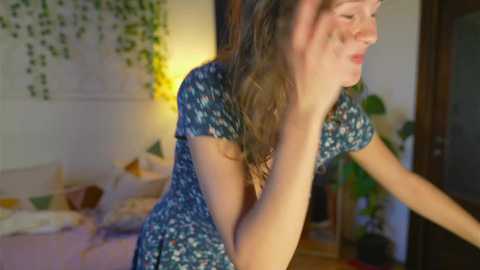 Media: Video of a smiling young woman with fair skin, brown hair, and wearing a blue floral dress, standing indoors near a potted plant, a bed, and hanging greenery.