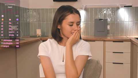 Media: Video of a young woman with shoulder-length dark hair, wearing a white shirt, sitting in a modern kitchen with tiled backsplash and wooden countertops. She covers her face with her hands, appearing upset.