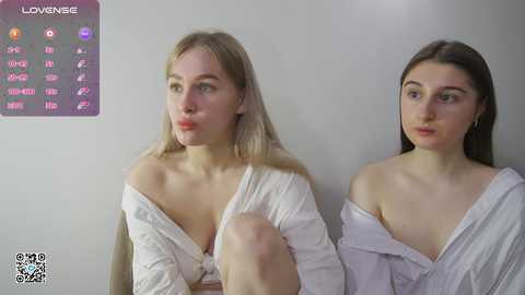 Media: Video of two young women with fair skin, blonde and brunette hair, wearing off-shoulder white tops, sitting side by side, looking serious.