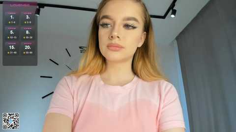 Media: Video of a young woman with fair skin, light brown hair, and large, bold eyelashes, wearing a pink t-shirt, standing in a modern room with a clock and calendar on the wall.