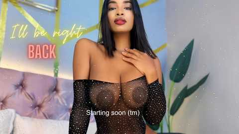 Media: Video of a confident, dark-skinned woman with straight black hair, wearing a sheer black top revealing her breasts, standing in a modern, brightly lit room with green accents and a \"BACK\" sign.