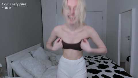 Media: Video of a slender, fair-skinned blonde woman with messy hair, wearing a brown strapless bra and white pants, standing in a minimalistic bedroom with a white bed and black-and-white cow-patterned blanket.