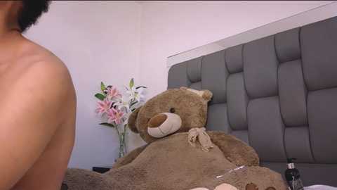 Media: Video of a large, plush teddy bear in a gray tufted headboard bedroom, with a man's bare back visible, and a vase of pink and white lilies on a white wall.