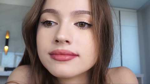 Media: Video of a young Caucasian woman with light skin, straight brown hair, and brown eyes. She wears natural makeup with light pink lips and subtle eye makeup. Background shows a modern bathroom with a white cabinet and a wall-mounted lamp.