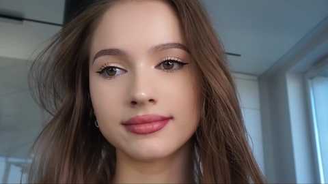 Media: Video of a young woman with fair skin, long brown hair, and light makeup, including nude lipstick and winged eyeliner. She has a calm expression and is indoors with a blurred background.