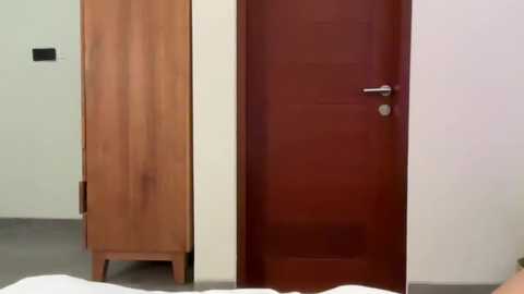 Media: A video of a bedroom with a closed, dark brown wooden door and a light wooden wardrobe standing side by side against a plain white wall. A white bed is partially visible in the foreground.