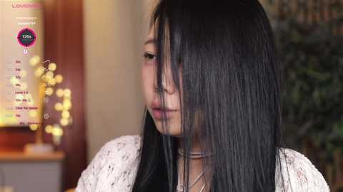 Media: A video of a young woman with long black hair, wearing a white floral top, looking down thoughtfully. Background shows a blurred, warm-toned room with a digital display.