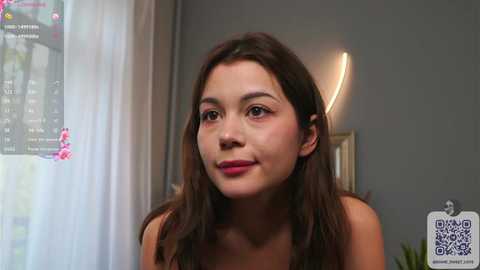 Media: Video of a young woman with long dark hair, wearing a sleeveless top, smiling in a modern room with a gray wall, a lamp, and a window with sheer curtains.