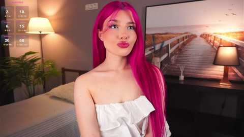 Media: Video of a young woman with long, vibrant pink hair and pink lipstick, wearing a white off-the-shoulder top, standing in a modern bedroom with a bed, potted plant, and framed picture of a bridge.