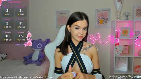 Media: A video of a young woman with long black hair, wearing a revealing, black-ribboned top, sitting on a plush chair in a pink-themed room with a neon sign and plush toys.