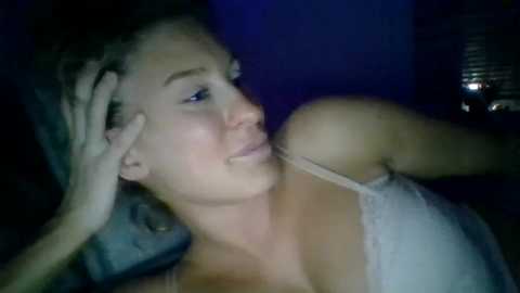 Media: Video of a fair-skinned woman with short, curly hair, wearing a white lace bra, lying on a bed. She appears relaxed, with her left hand on her forehead. Background is dimly lit with a blue hue, and blinds are partially closed.