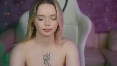 Media: Video of a topless young woman with long blonde hair, closed eyes, and a tattoo of a dragon on her chest. She sits on a white chair in a dimly lit room with purple curtains and a purple background.