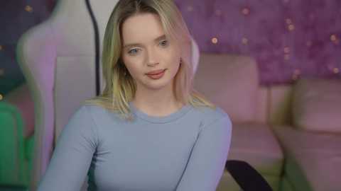 Media: Video of a young woman with shoulder-length blonde hair, wearing a light blue long-sleeve top, seated indoors against a backdrop of purple and green lights and a tufted white couch.