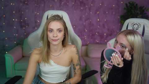 Media: Video of two young women in a gaming room; one with a white off-the-shoulder crop top, blonde hair, tattoos, and a nose ring; the other, also blonde, wearing a black top, holding a pink headset.