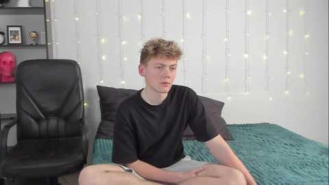 Media: Video of a young, fair-skinned, curly-haired man in a black T-shirt and white shorts, sitting cross-legged on a teal bed with a black chair and white string lights in the background.