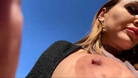 Media: Video of a topless woman with fair skin and blonde hair, wearing large pearl earrings, against a bright blue sky, focusing on her large, exposed breasts.