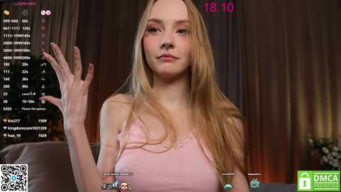 Media: Video of a young woman with long blonde hair and fair skin, wearing a pink camisole, making a peace sign. Background includes a dimly lit room with streaming platform logos.