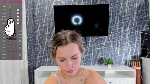 Media: A video of a young woman with light brown hair, wearing a beige top, sits in a room with a white wall, black TV, and a penguin-themed poster.