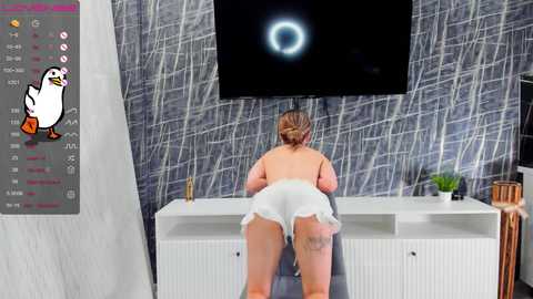 Media: Video of a nude woman with blonde hair, wearing only a white dress, bending over a white desk in a modern, minimalist room with gray wallpaper and a flat-screen TV.